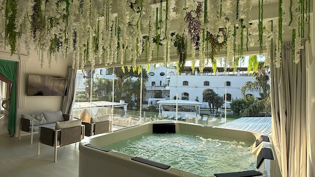 A modern indoor pool area with hanging floral decorations, a seating area with a sofa and chairs, and a view of the surrounding buildings. - Hotel Imperiale (Terzigno)