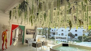 A spacious room with a high ceiling decorated with hanging plants, a pool area, and a view of a building outside. - Hotel Imperiale (Terzigno)