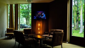 A room with a TV mounted on the wall, a table with chairs, and large windows with a view of trees and a car outside. - Hotel de Elderschans (Aardenburg)