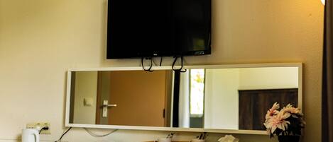 Une chambre d’hôtel équipée d’un bureau, d’un téléviseur à écran plat fixé au-dessus d’un grand miroir, d’une plante en pot et d’une prise électrique murale.