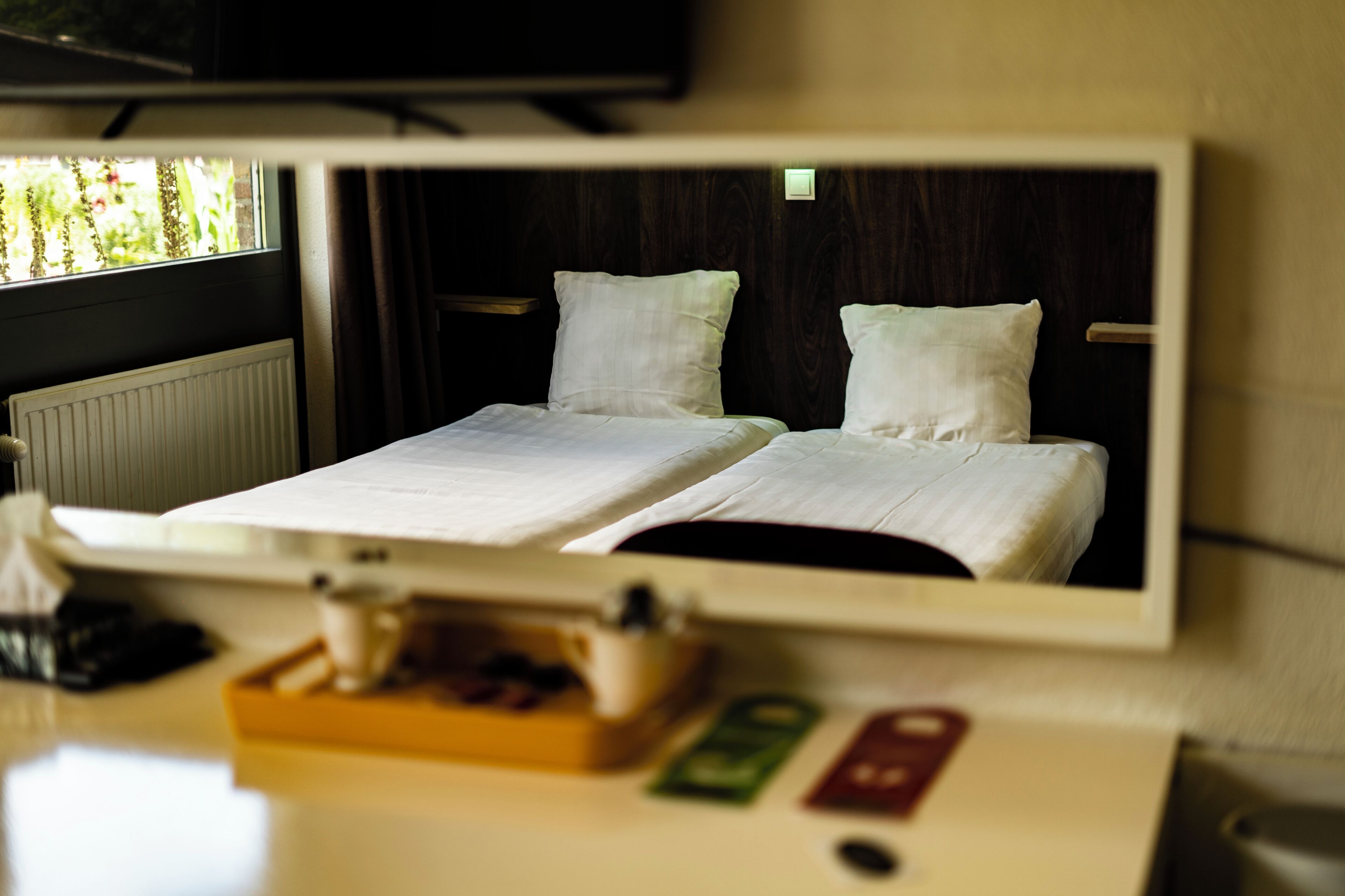 A hotel room with a bed, a bedside table with a cup and a tray, and a mirror reflecting the bed.