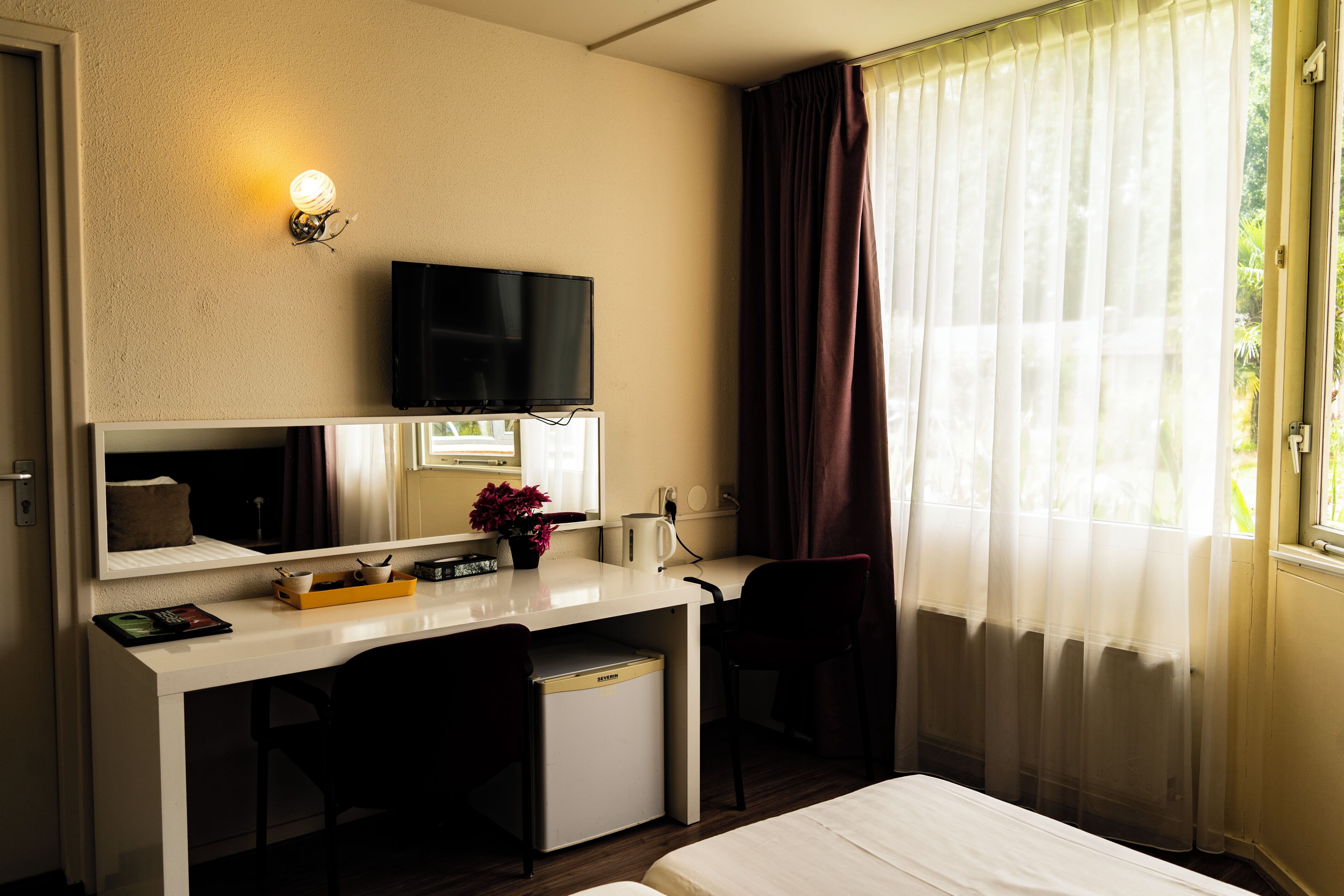 Habitación de hotel con cama, escritorio con televisor, silla, mueble pequeño y ventana con cortinas.