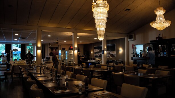 A restaurant with long communal tables, high ceilings, and chandeliers. - Hotel de Elderschans (Aardenburg)