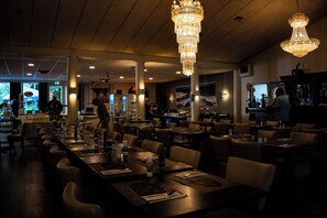 A restaurant with long communal tables, high ceilings, and chandeliers. - Hotel de Elderschans (Aardenburg)