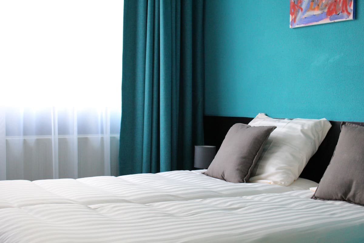 Una cama bien hecha con ropa de cama blanca y gris, cortinas color verde azulado y un cuadro en la pared.