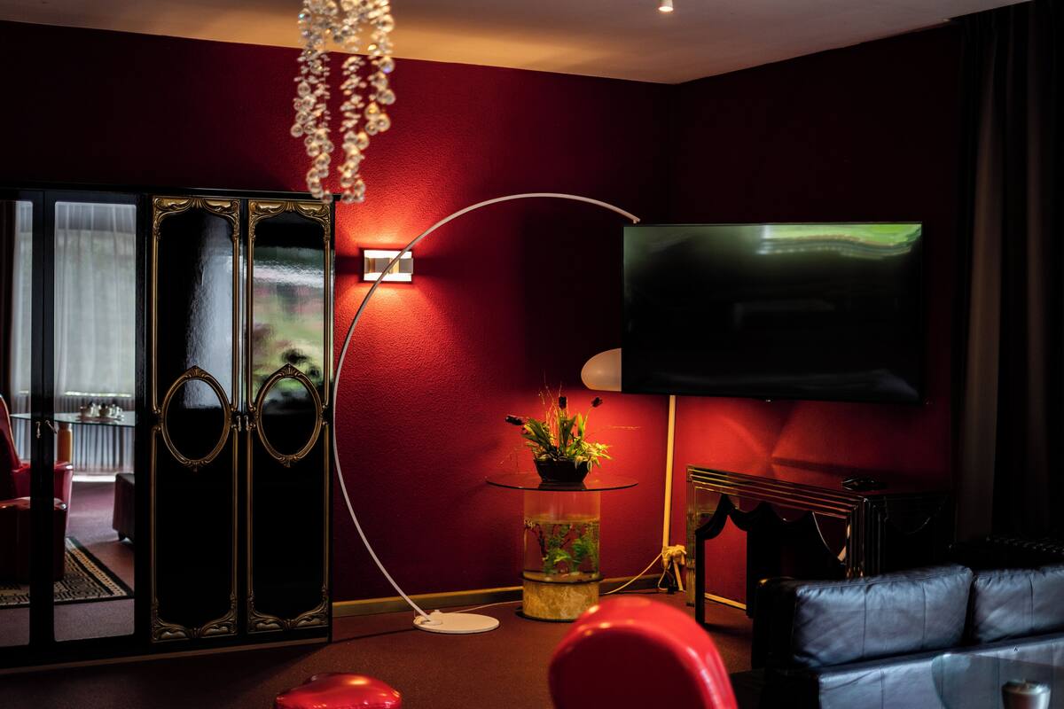 Una sala de estar con televisor, pared roja, mesa de vidrio con planta, sofá negro y lámpara decorativa.