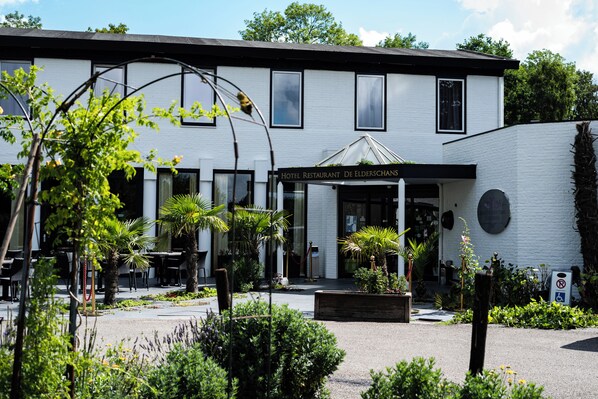 Un hôtel à la façade blanche, doté d’un espace extérieur couvert avec des sièges et d’une enseigne indiquant qu’il s’agit d’un restaurant.