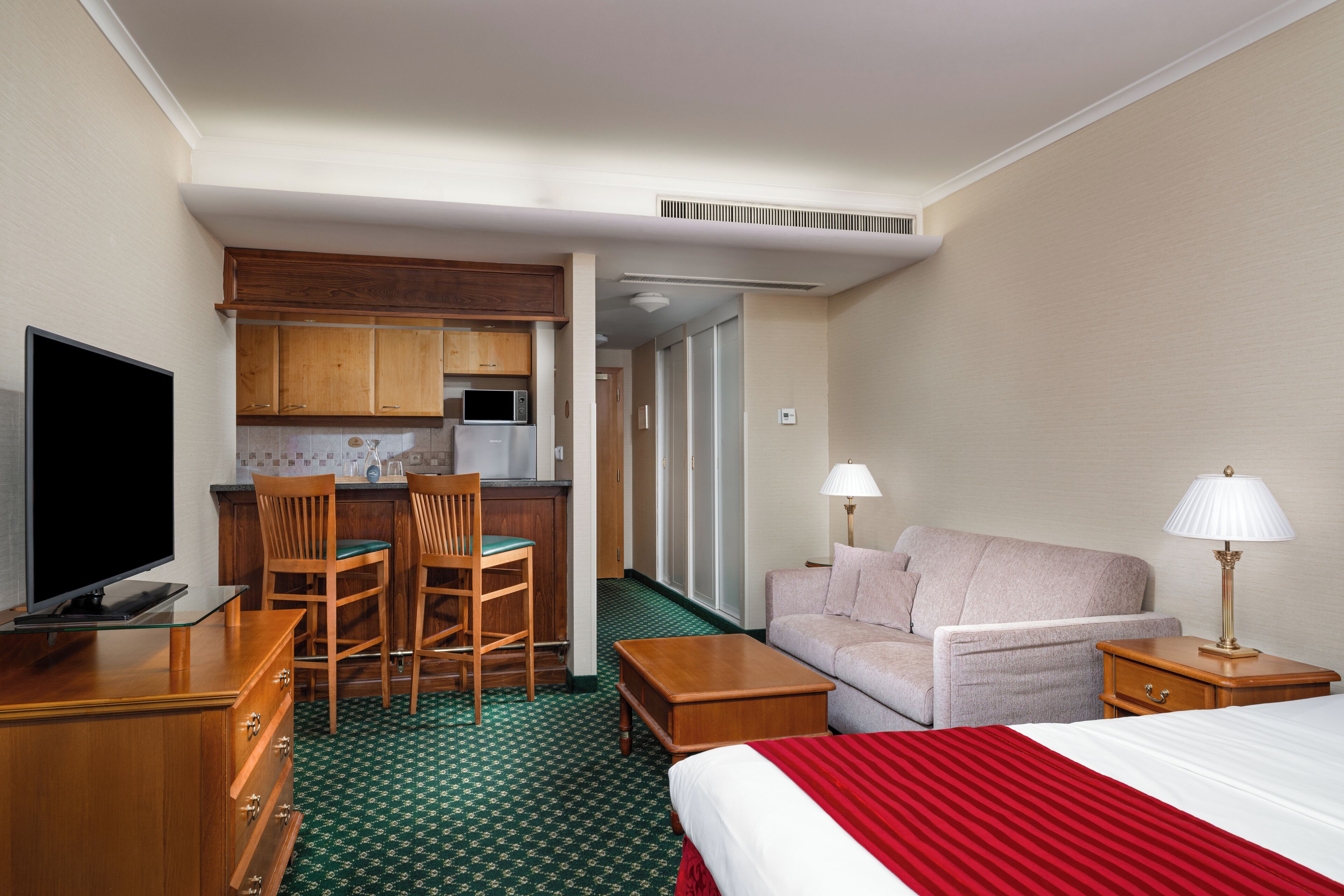 A hotel room with a bed, a sofa, a desk with a chair, a television, a kitchenette, and a bathroom.