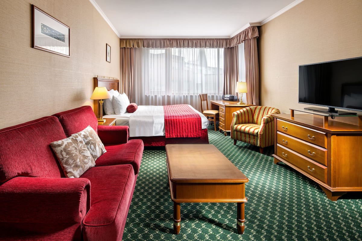 Een hotelkamer met een rode bank, een houten salontafel, een televisie, een bureau met een lamp en een groot raam met gordijnen.