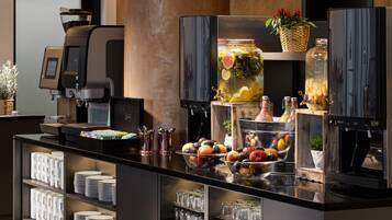 Disposición de un bufé con una máquina de café, fuentes de fruta y un frasco grande de verduras encurtidas.