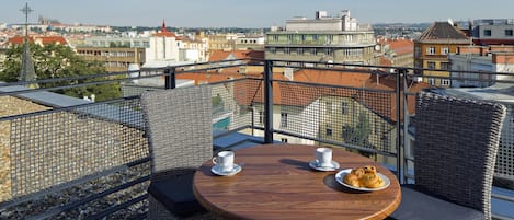 Una terraza en la azotea con sillas de mimbre y una mesa de madera, dos tazas blancas y un plato de pasteles.