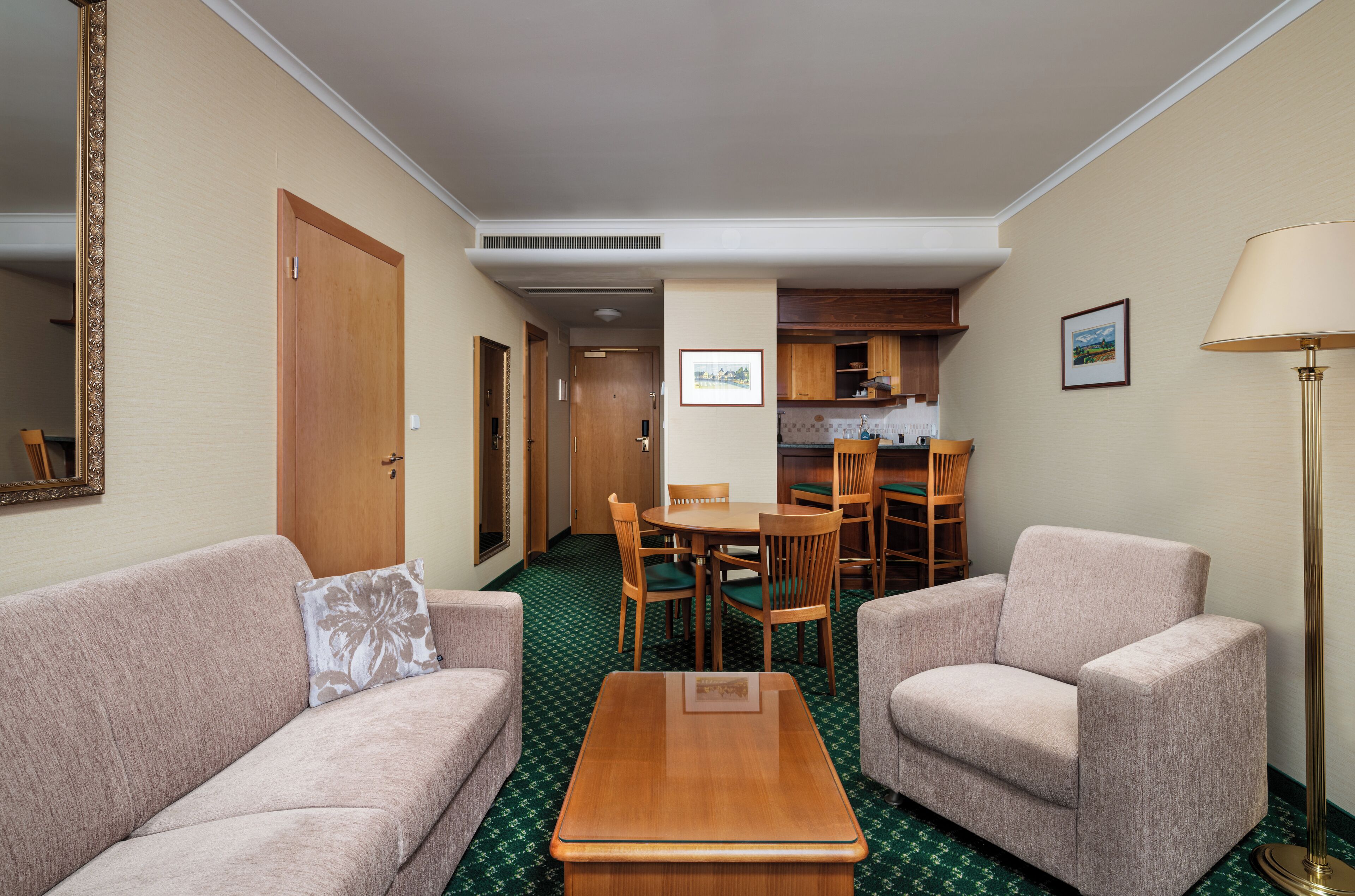 A hotel room with a green patterned carpet, two sofas, a wooden coffee table, a dining table with chairs, and a kitchenette.