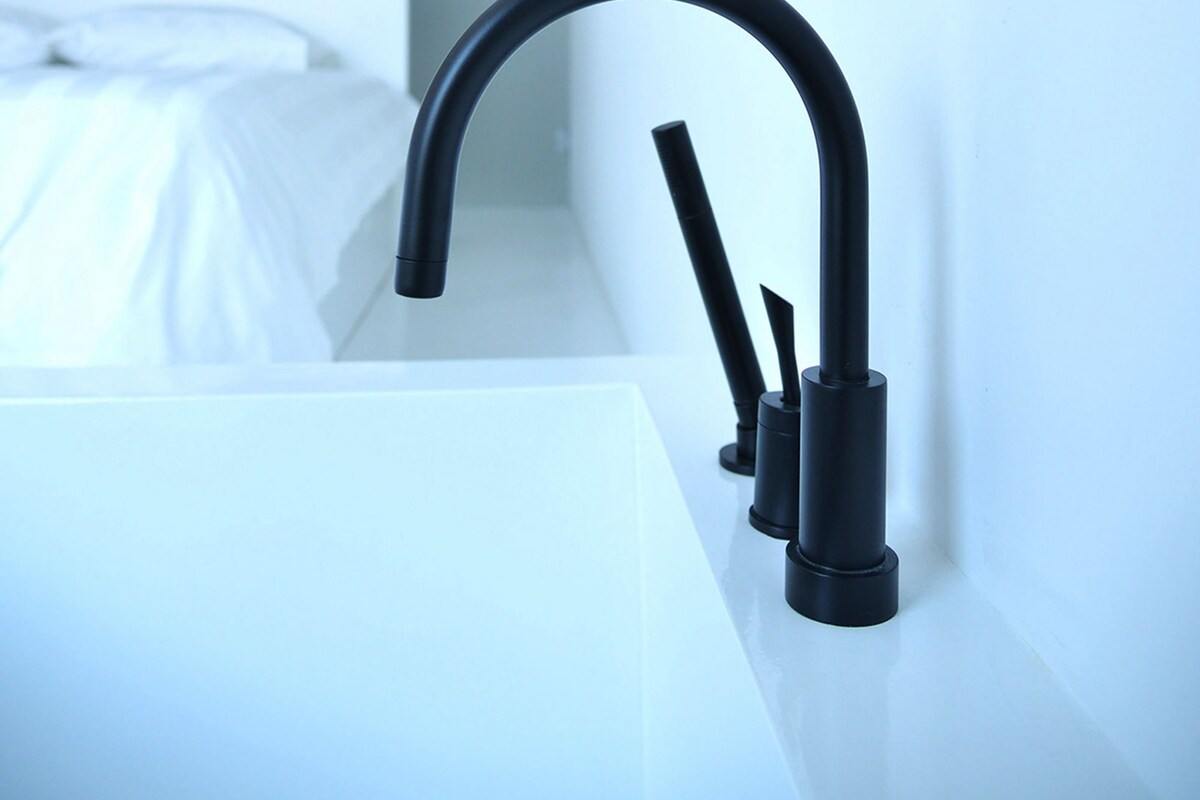 A modern bathroom with a white sink, black faucet, and a bed with white bedding in the background.