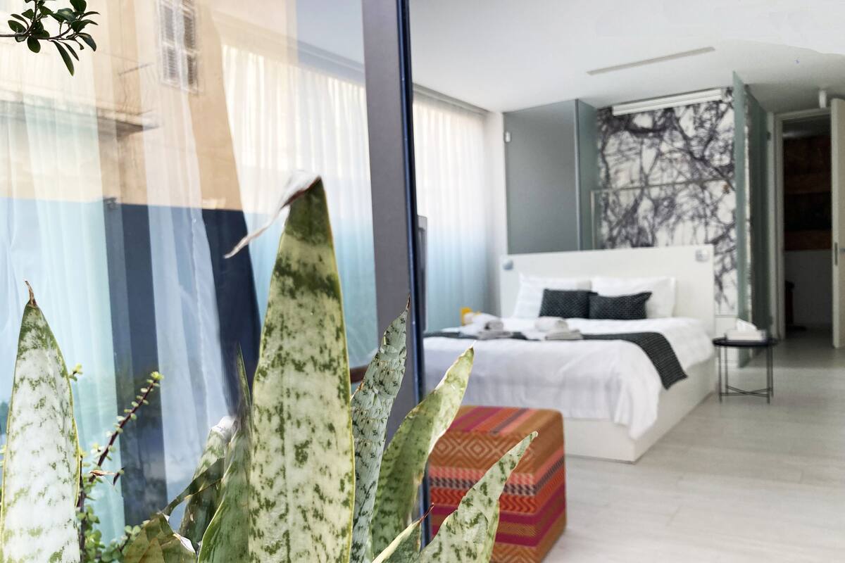 A modern hotel room with a large bed, a bedside table, and a window with sheer curtains.