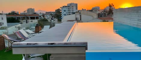 A rooftop pool area with lounge chairs and a view of the sunset over a cityscape.