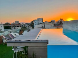 Área de piscina no terraço com espreguiçadeiras e vista para o pôr do sol sobre a paisagem urbana.