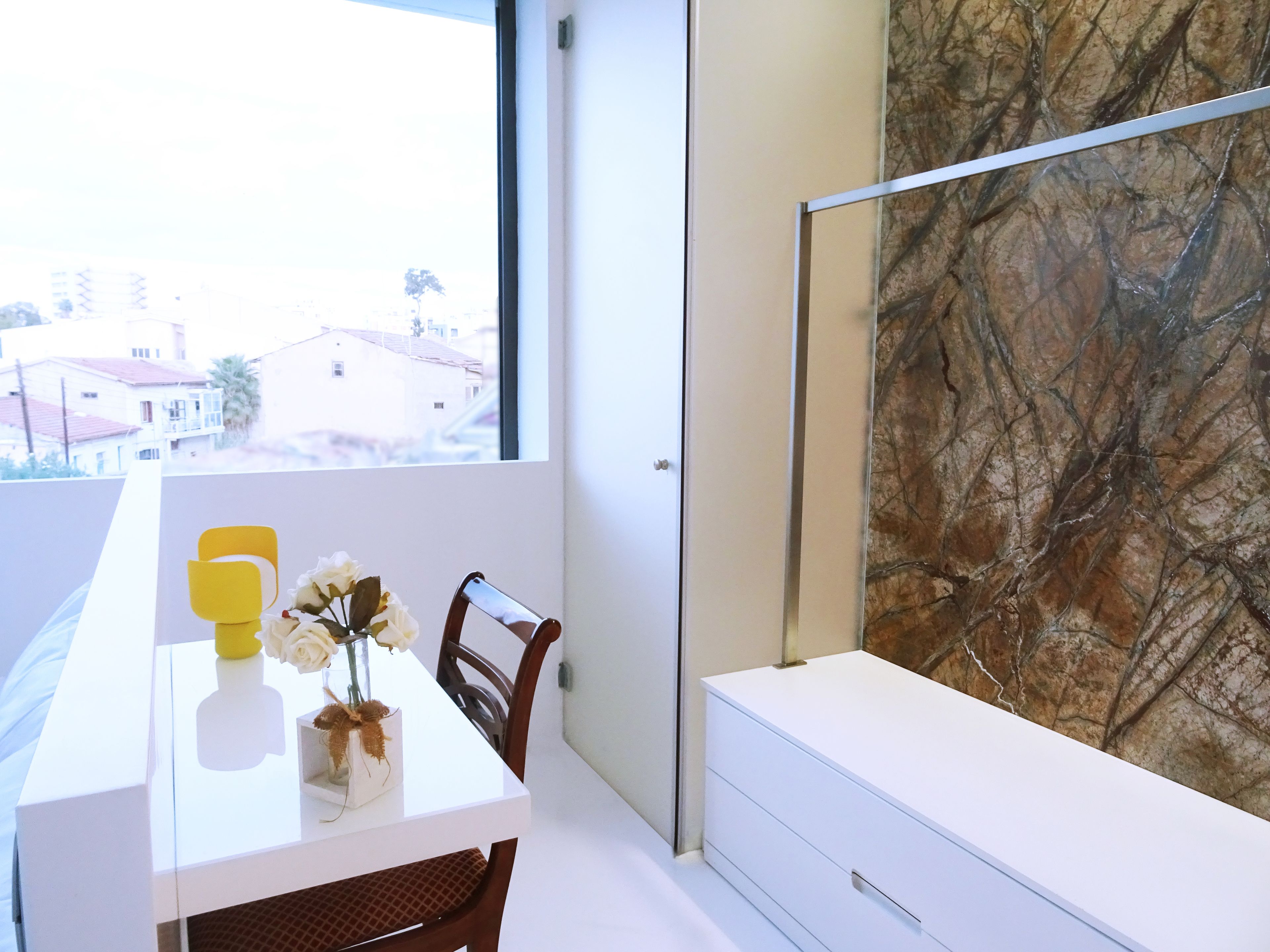 A modern room with a white table, a vase with flowers, a yellow cup, and a view of buildings outside a window.