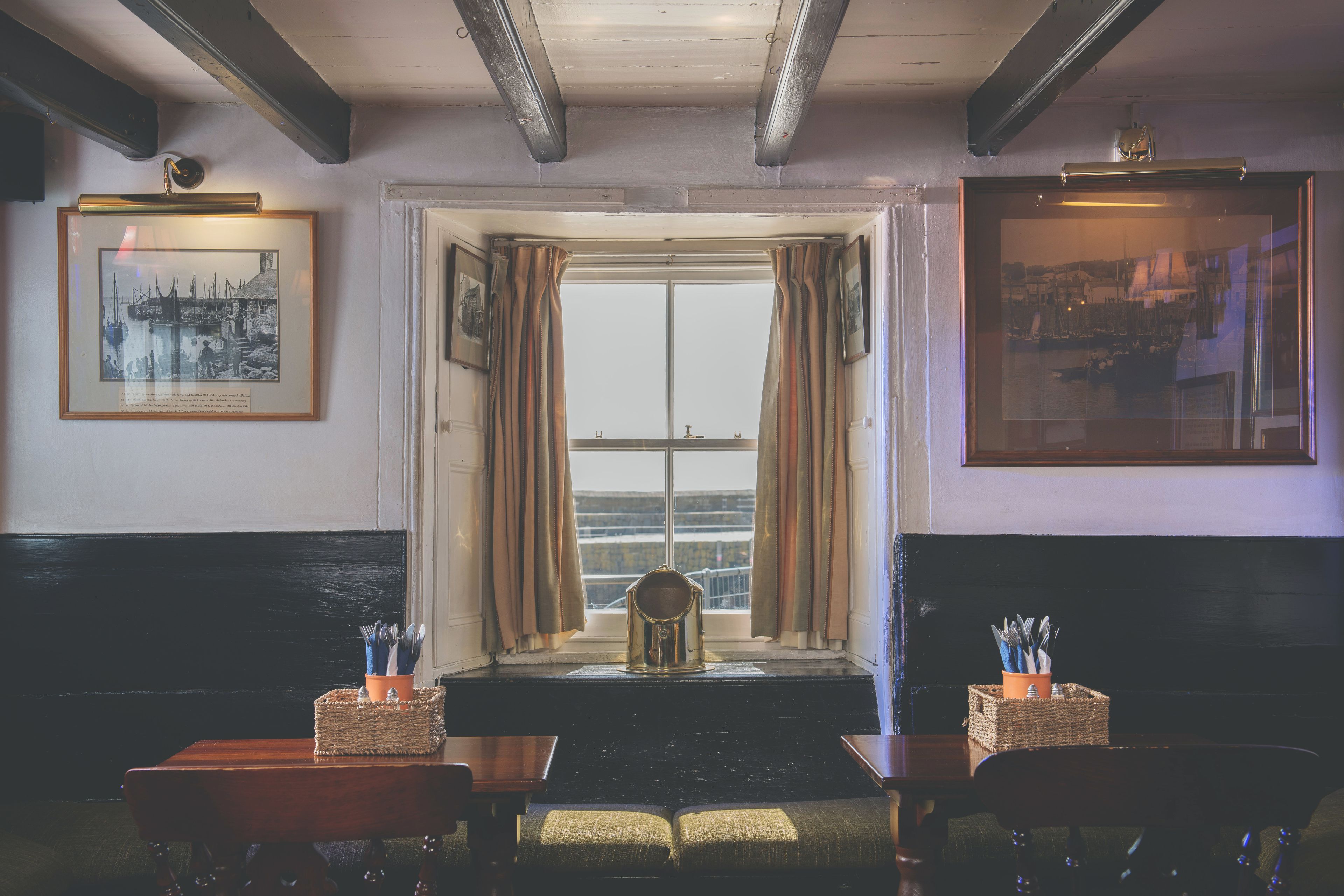 A restaurant interior with a view through a large window, a wooden bench, a table with a basket of cutlery, and framed pictures on the walls.