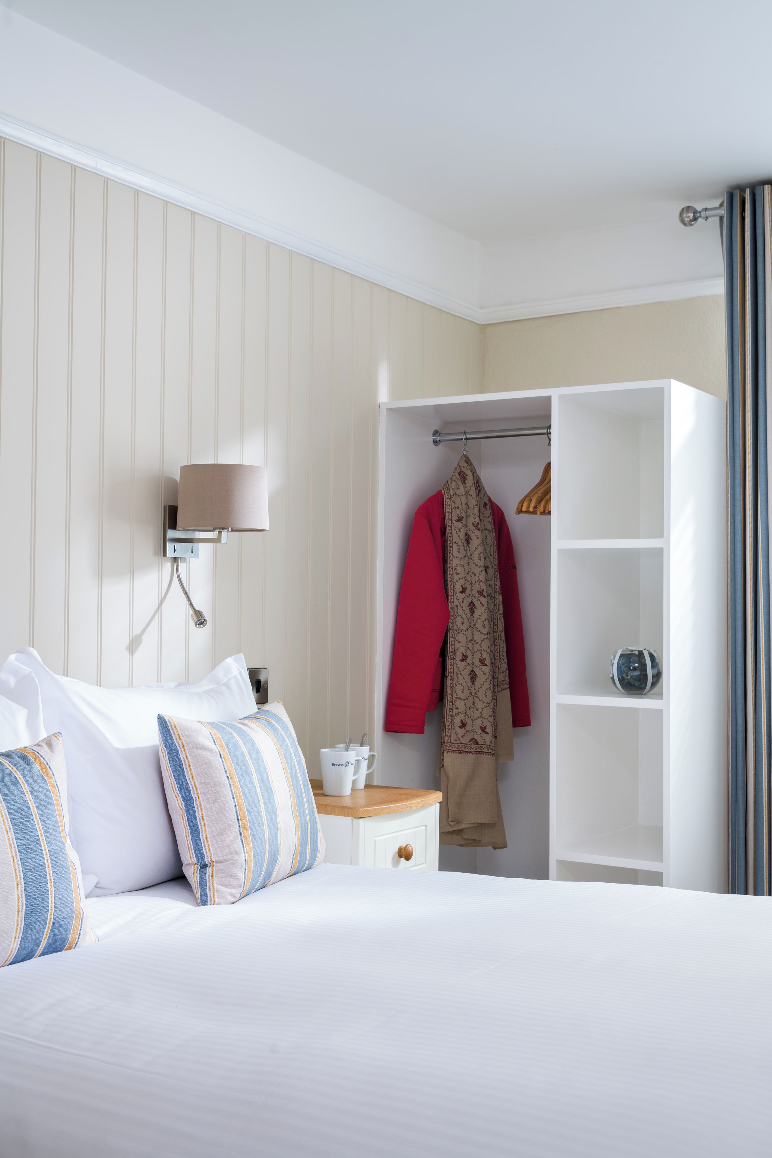 A hotel room with a bed, a wardrobe, a bedside table with a lamp, and a shelf with decorative items.