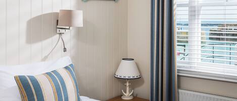 A hotel room with a bed, a nightstand with a lamp, a wall-mounted sign reading 'BEACH', and a window with blinds.