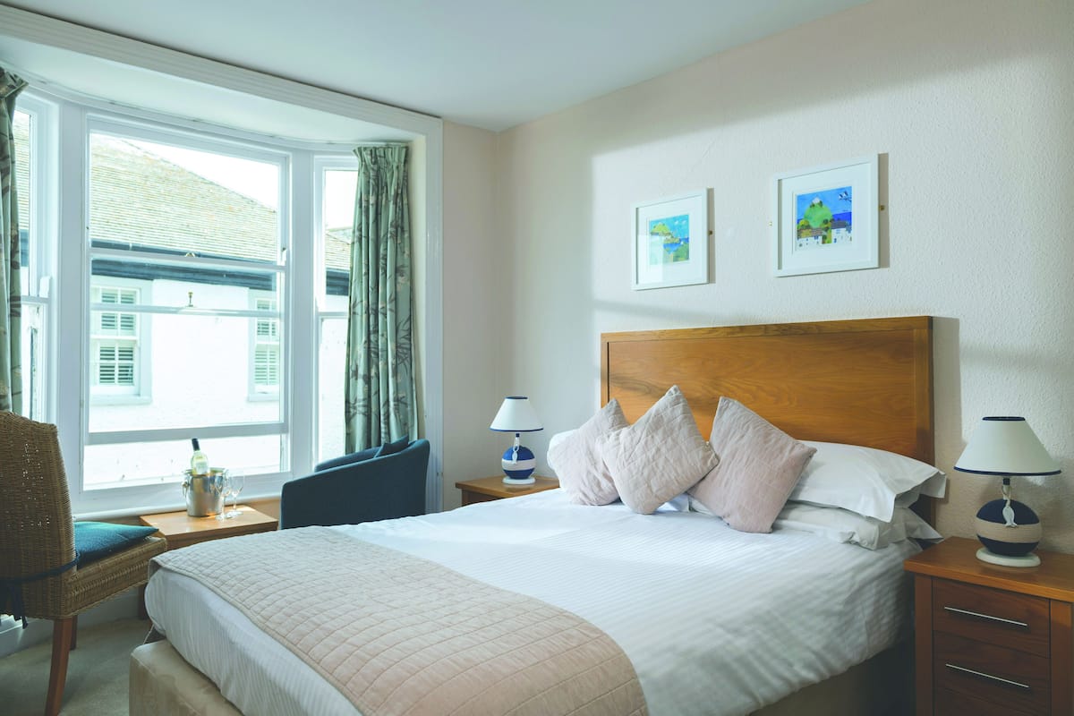 Quarto de hotel com uma cama grande, duas mesas de cabeceira, uma cadeira e uma janela com cortinas.