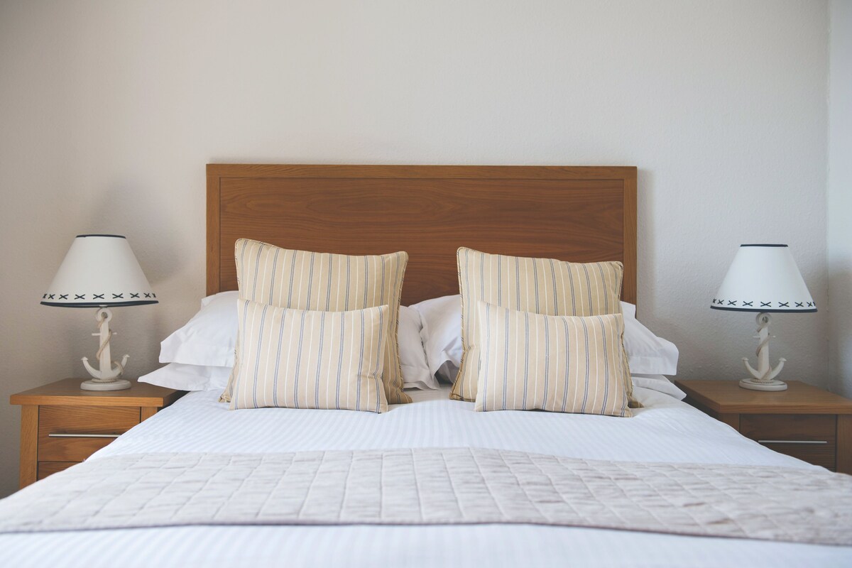 Un lit bien fait, agrémenté d’oreillers à rayures et d’une couverture matelassée, avec deux tables de chevet de chaque côté, sur lesquelles sont posées des lampes.