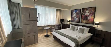 Habitación de hotel moderna con una cama grande, un escritorio, una silla y una mesita pequeña. Hay dos cuadros en la pared y una ventana con cortinas.