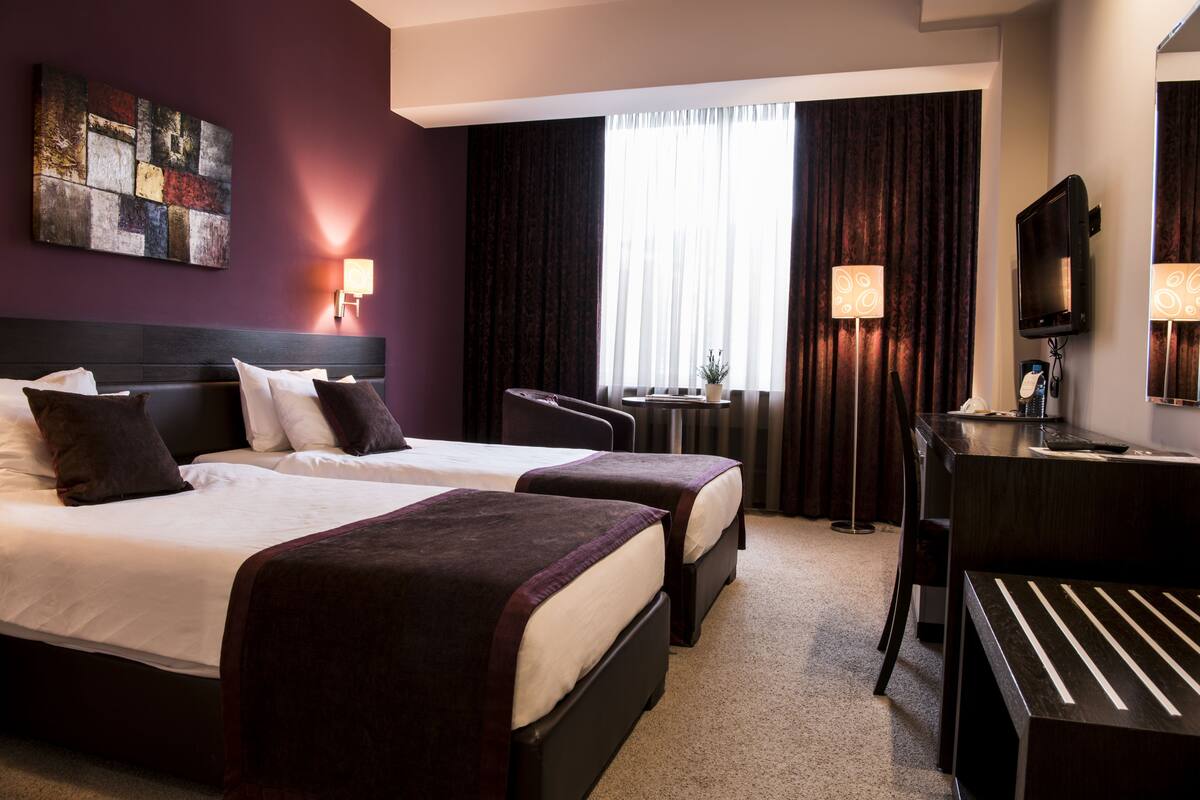 A hotel room with two beds, a desk, a chair, a TV, and a window with curtains.