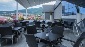 An outdoor seating area with tables and chairs, potted plants, and a view of the city and mountains. - Queen's Hotel (Skopje)