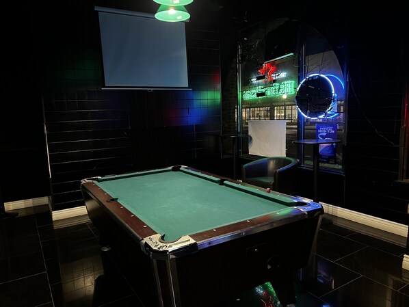 A pool table in a dimly lit room with a neon sign and a projector screen.