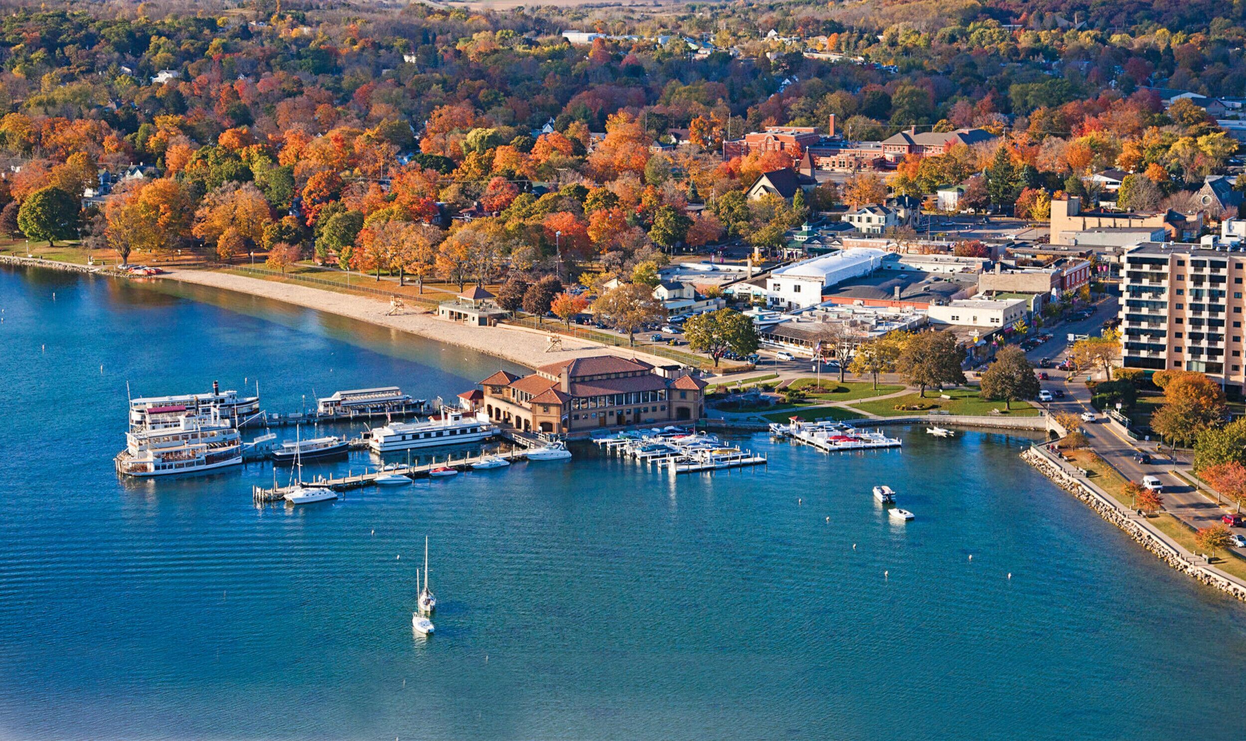 A waterfront marina with multiple boats docked, surrounded by autumn-colored trees and buildings.