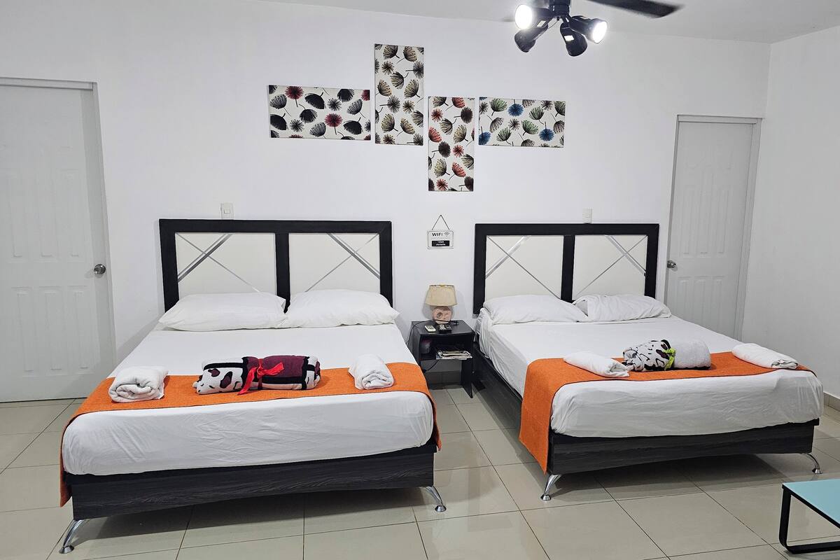 A hotel room with two beds, white bedding, orange throw blankets, a cross-patterned wall decoration, a ceiling fan, and a small table with a vase of flowers.