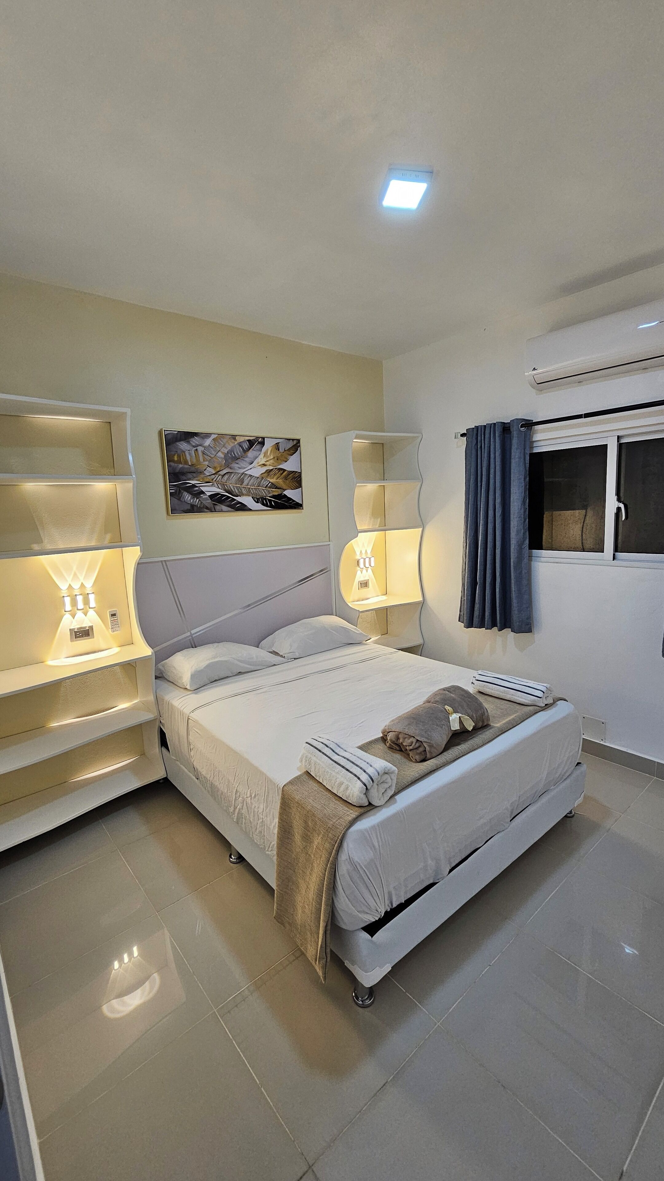 A modern hotel room with a large bed, bedside lamps, a wall-mounted air conditioner, and a window with curtains.