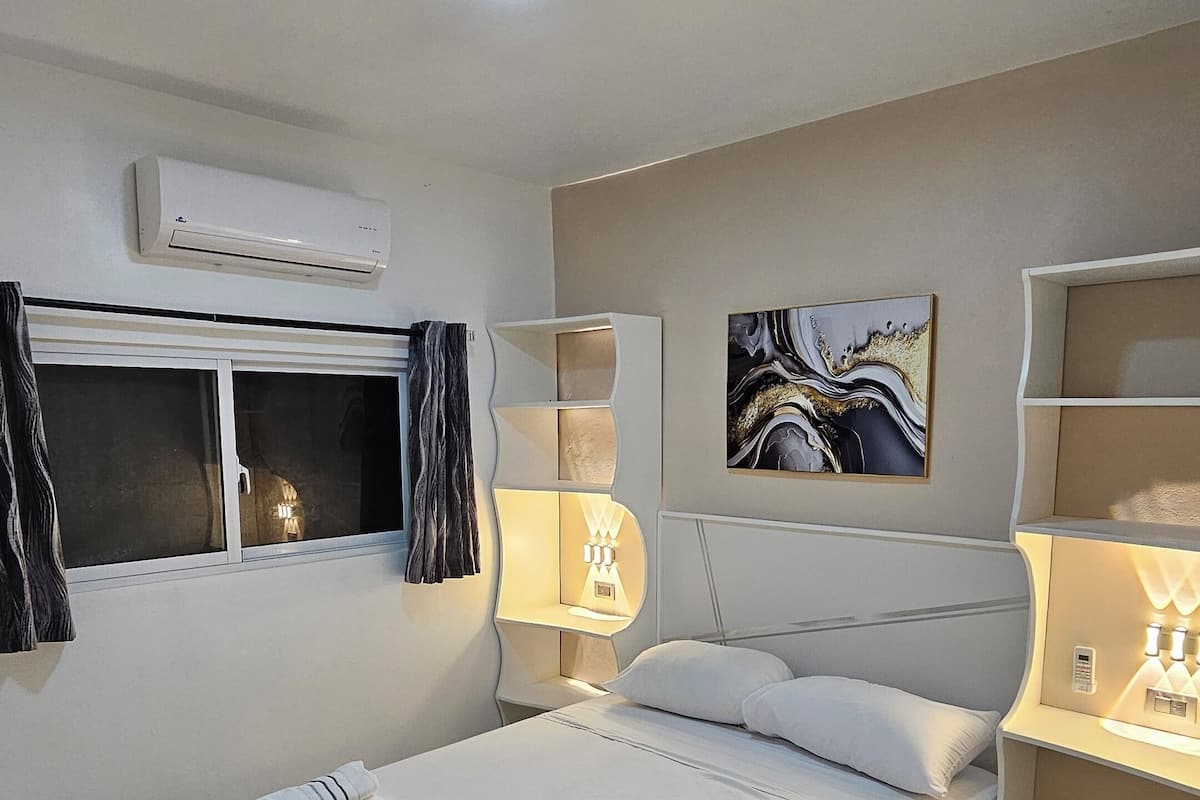 A modern bedroom with a large bed, bedside lamps, an air conditioner, and a window with curtains.
