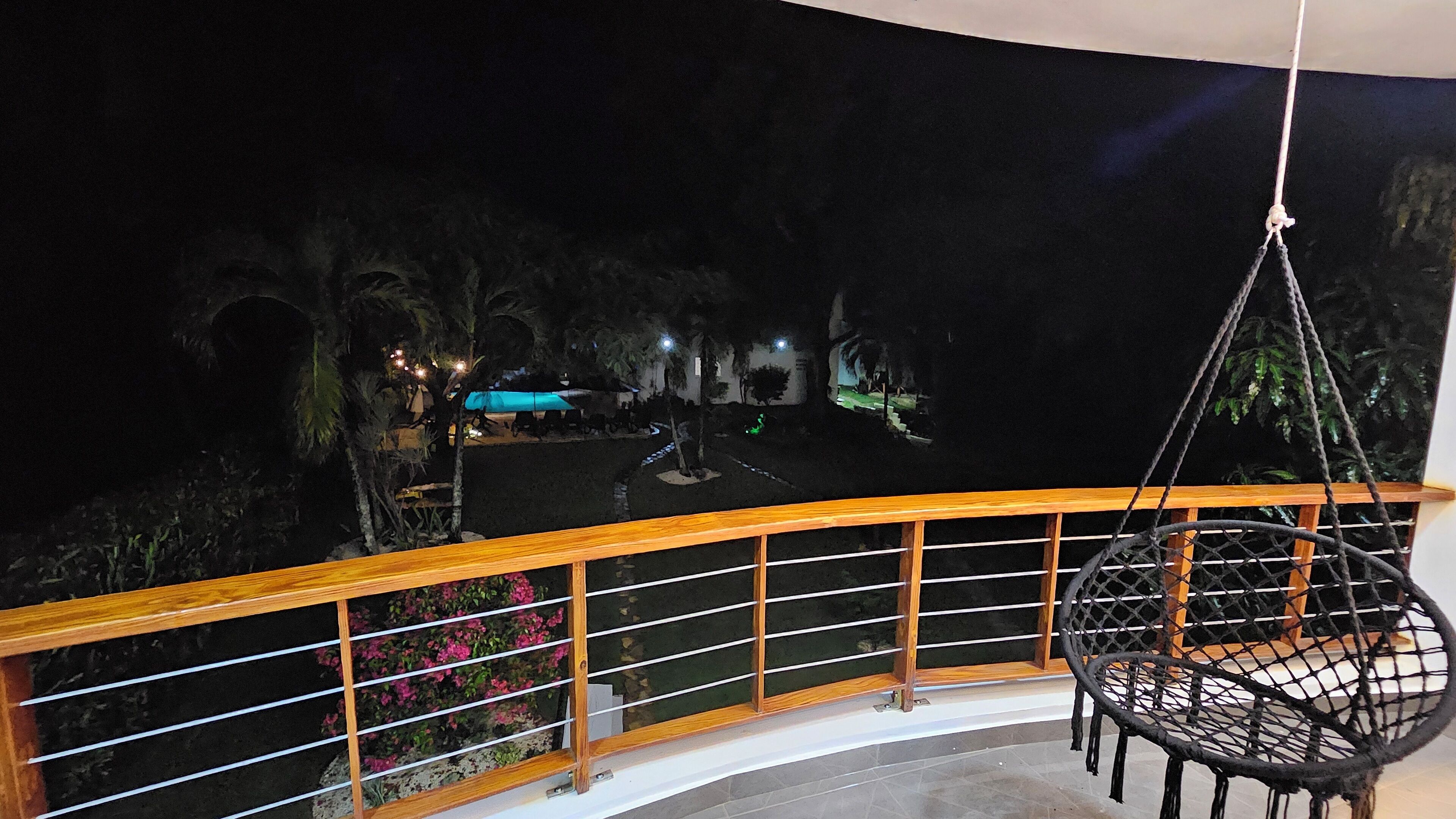 A balcony with a hanging chair, a railing, and a view of a pool area at night.