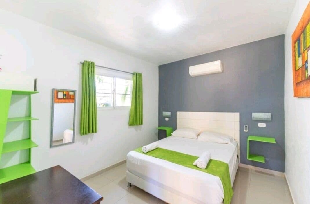 A hotel room with a bed, a desk, a mirror, and a window with green curtains.