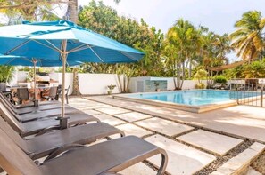 A poolside area with lounge chairs and a blue umbrella, surrounded by palm trees and a stone-tiled deck. - Villa Victoria (Sosúa)
