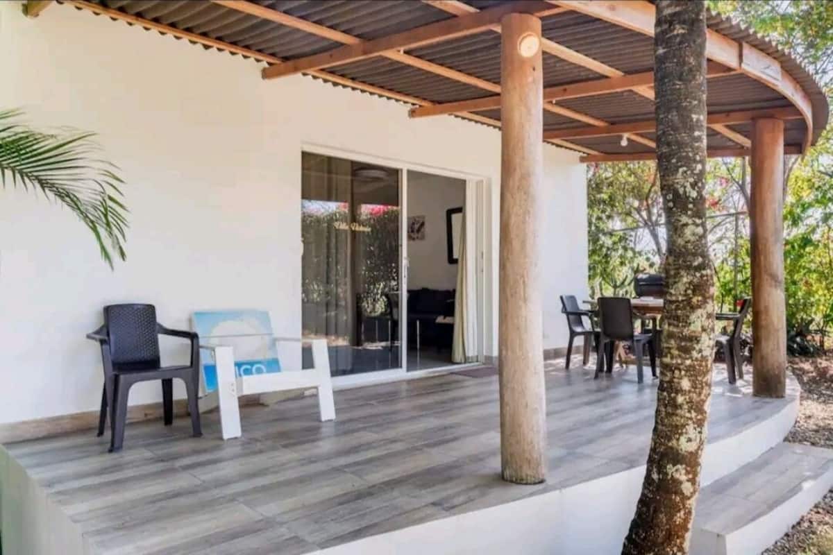 A covered outdoor area with a wooden pergola, a seating area with chairs and a table, and a glass door leading to an interior space.