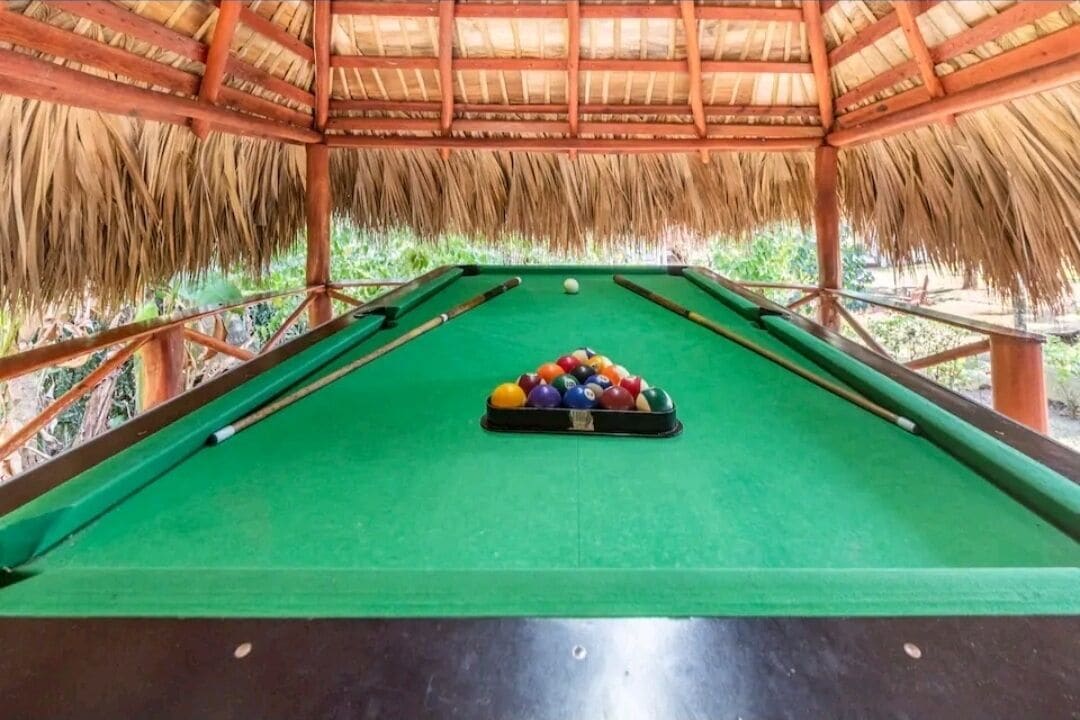 A pool table with a thatched roof overhead, surrounded by a wooden railing.