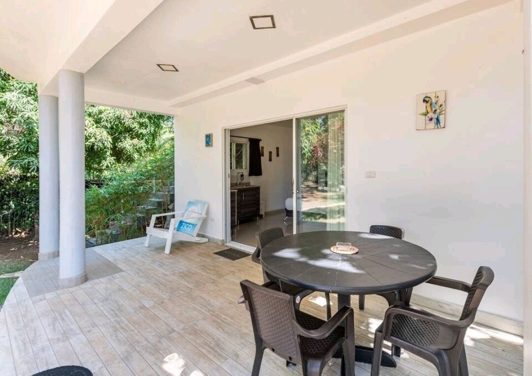 A modern outdoor dining area with a round table and chairs, a glass door leading to a kitchen, and a view of greenery.