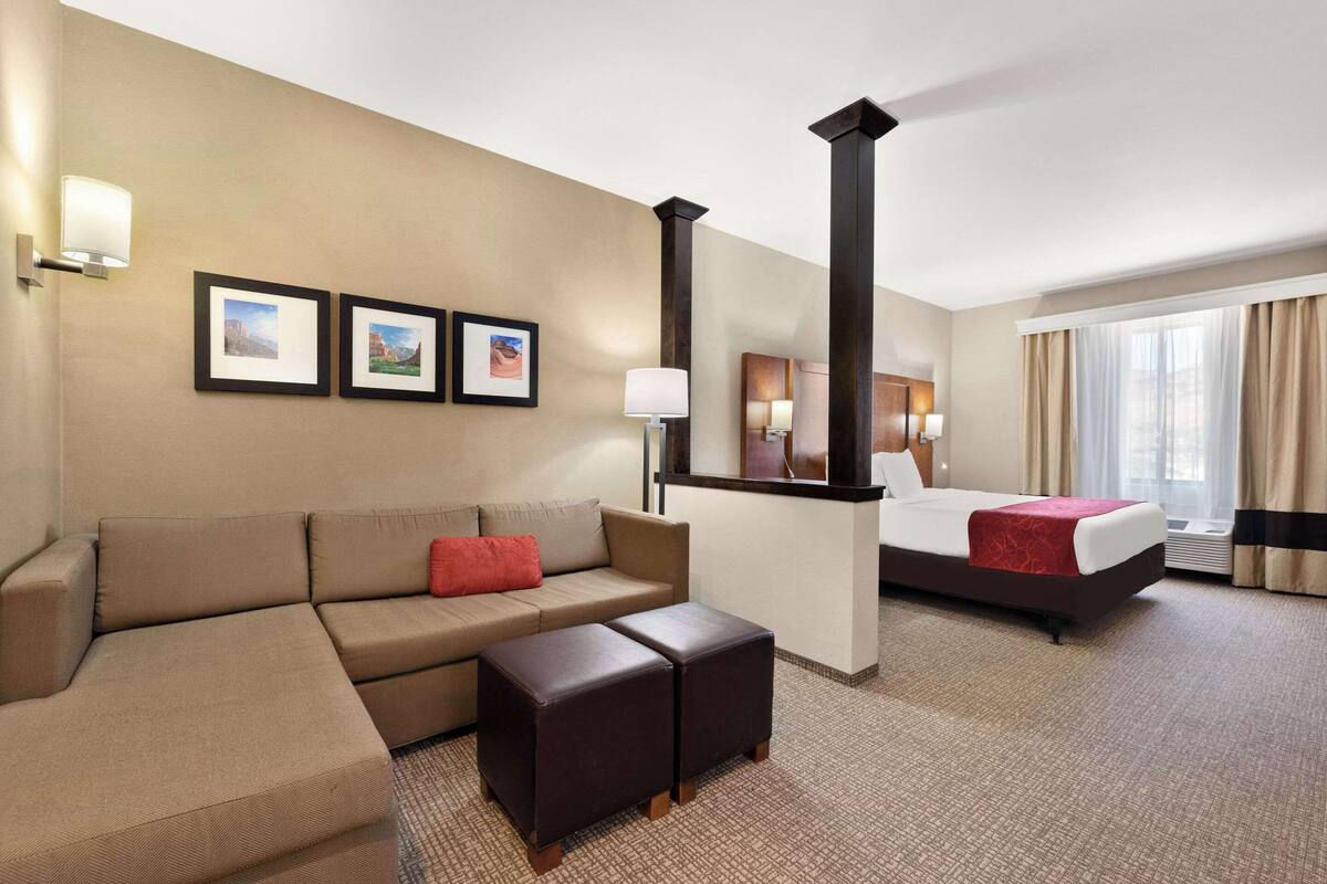 Ein Hotelzimmer mit Bett, einer Couch, einem Couchtisch und einem Schreibtisch. An der Wand hängen Bilder in Rahmen und ein Fenster mit Vorhängen.