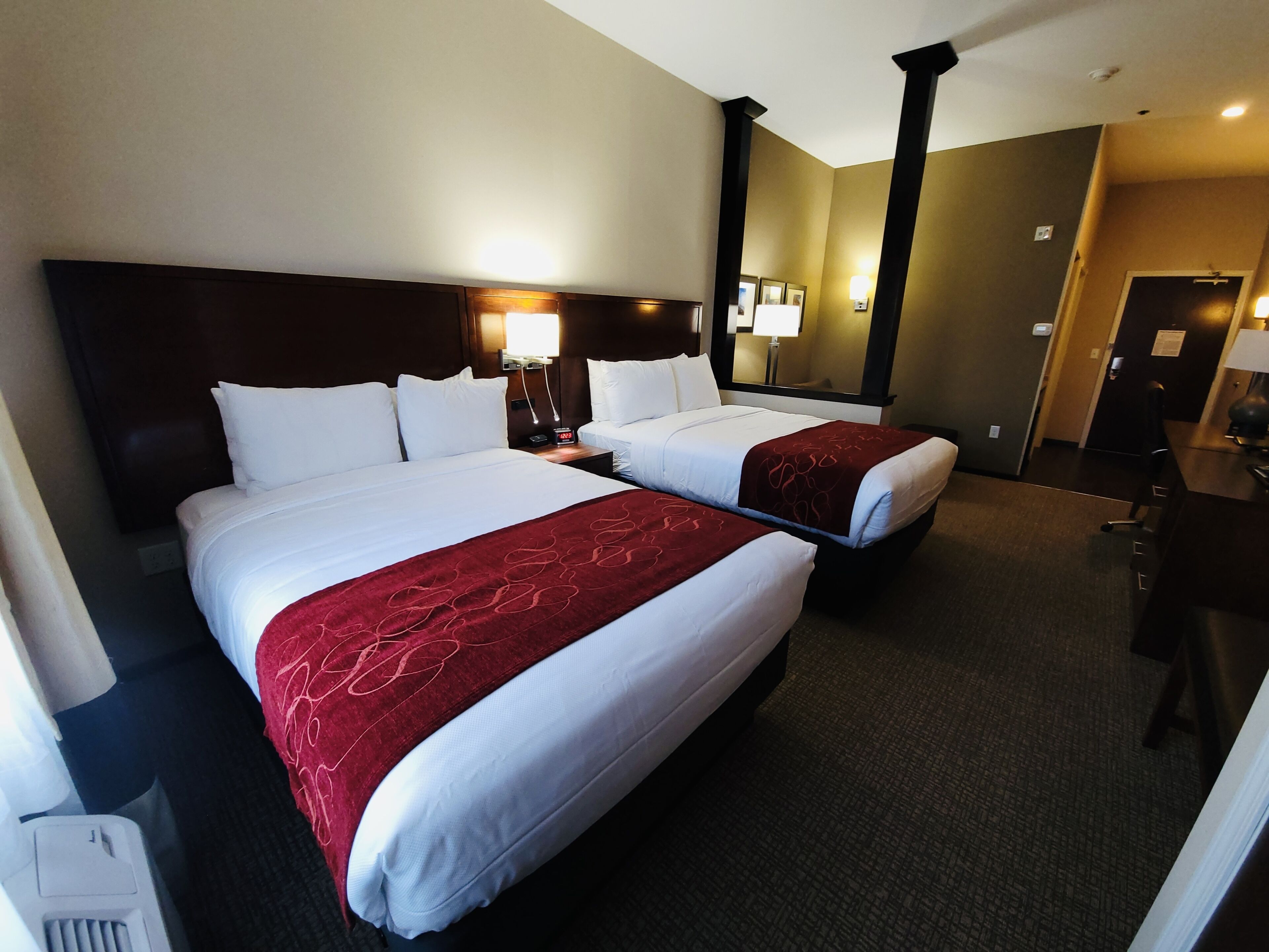 A hotel room with two beds, a dark headboard, bedside lamps, a mirror, and a desk with a chair.
