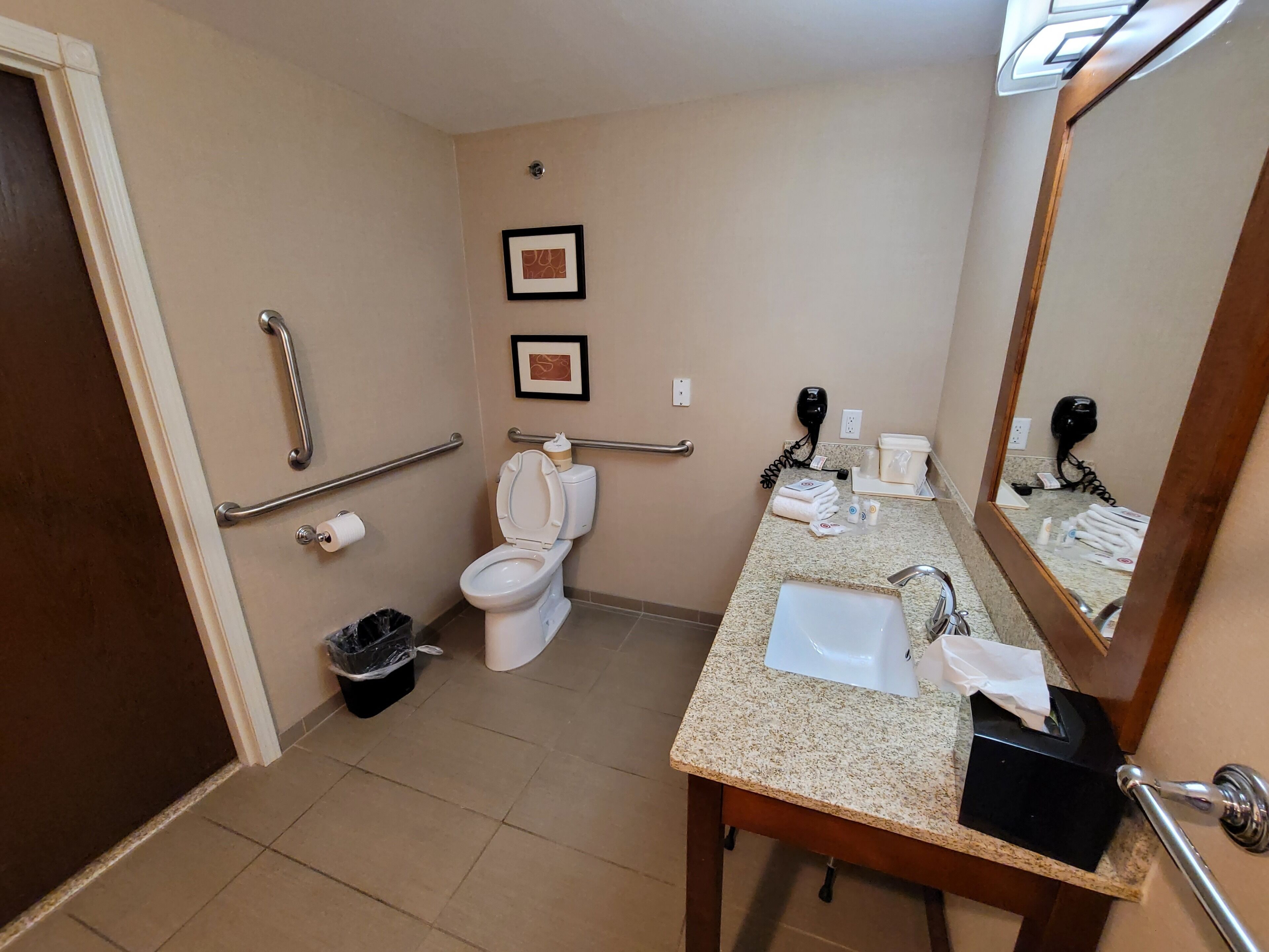 Une salle de bain d’hôtel standard, équipée de toilettes, d’un lavabo, d’un miroir, de barres d’appui et d’une poubelle.