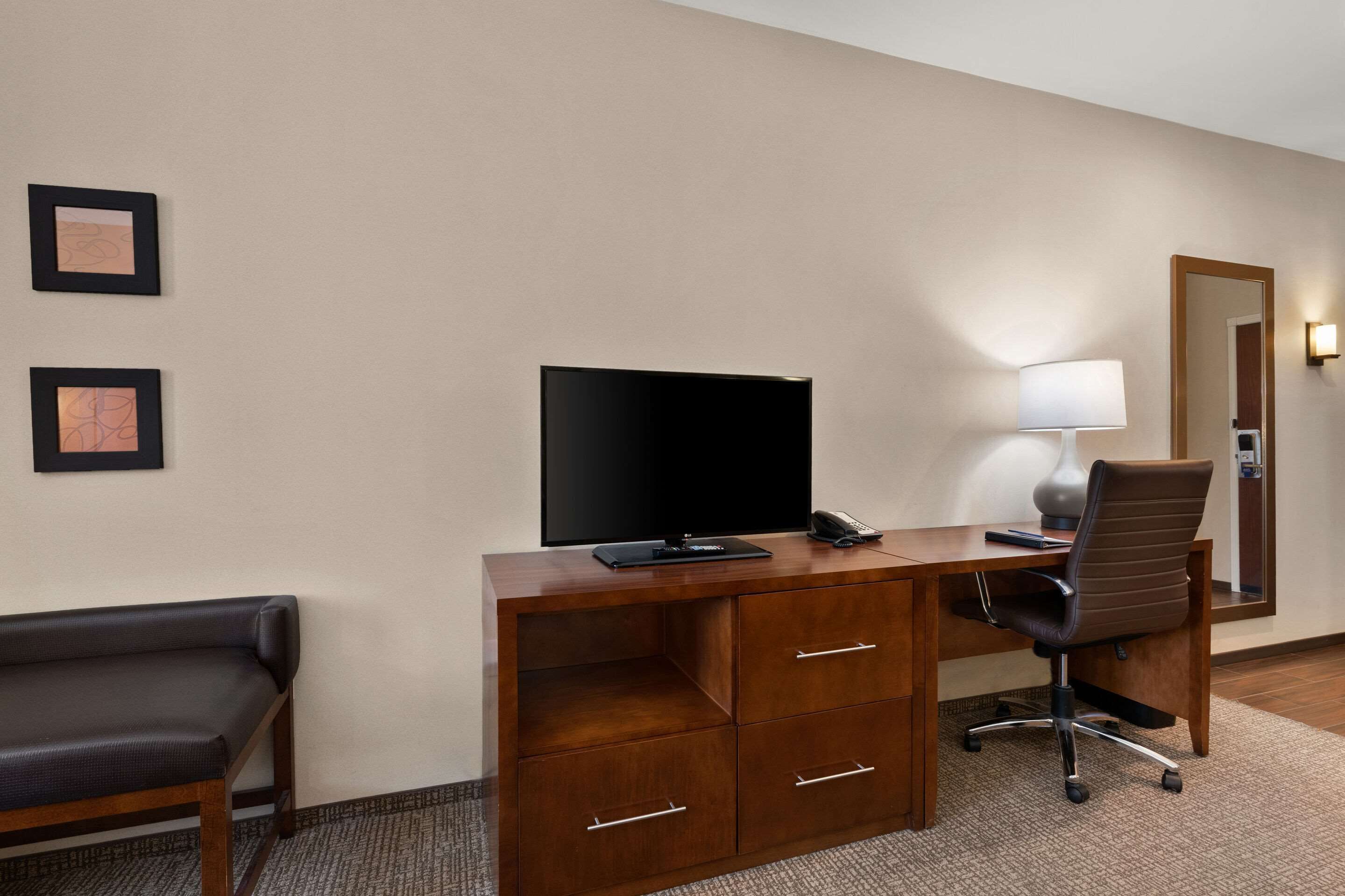A hotel room with a wooden desk, a flat-screen TV, a lamp, a chair, and a bench.