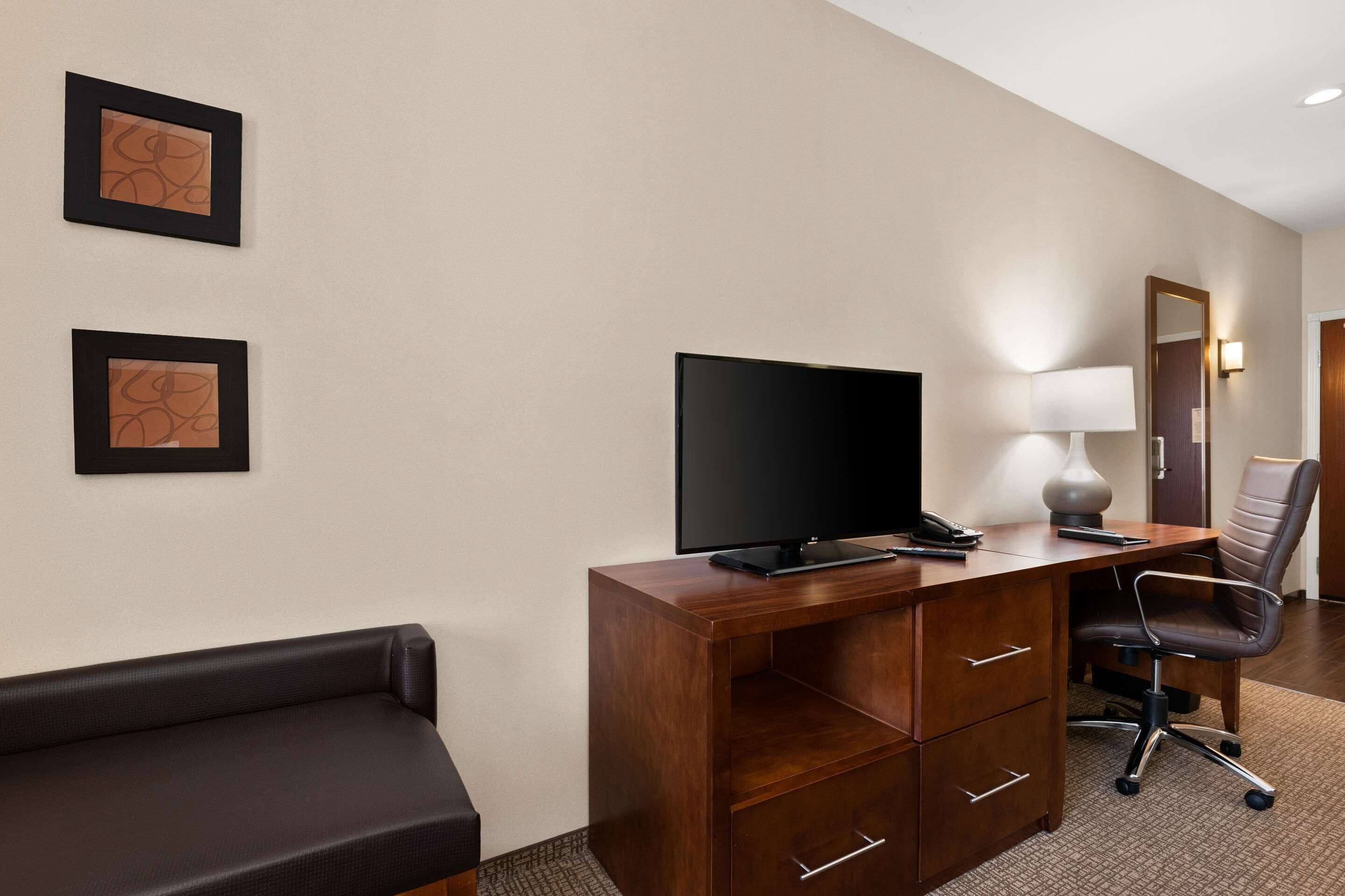 A hotel room with a wooden desk, a flat-screen TV, a lamp, a chair, and two framed pictures on the wall.