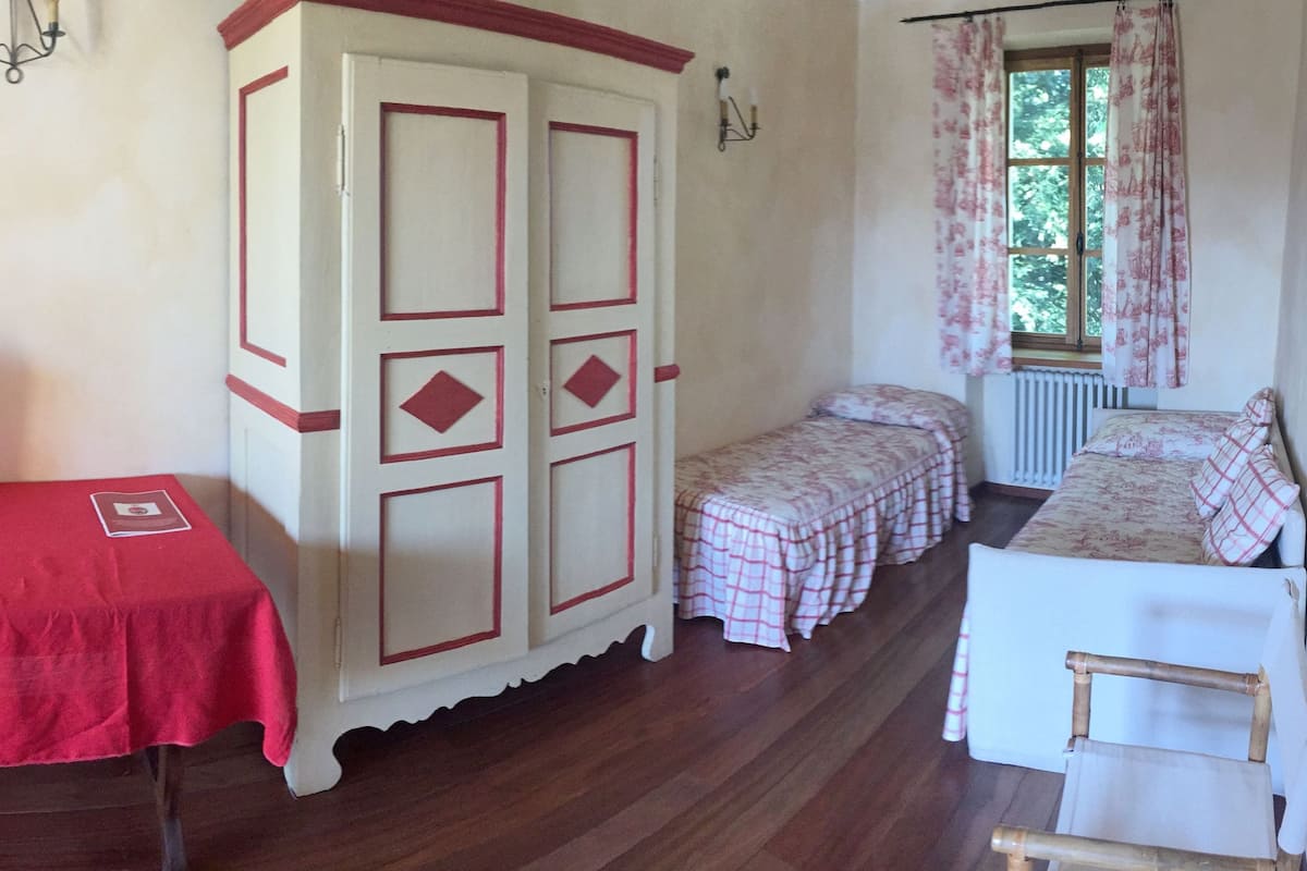 A room with a red table, a white cabinet with red trim, a bed with a floral bedspread, and a window with curtains.