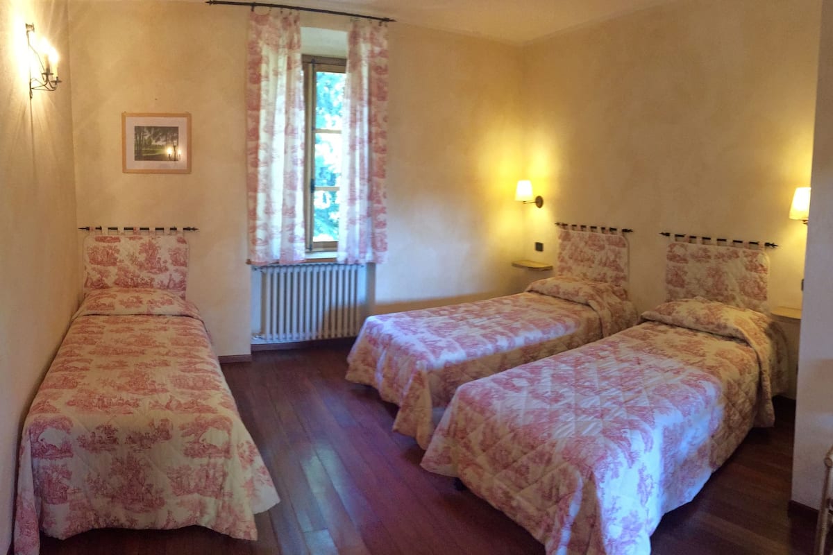 A room with two single beds, a radiator, a window with curtains, and wall-mounted lamps.