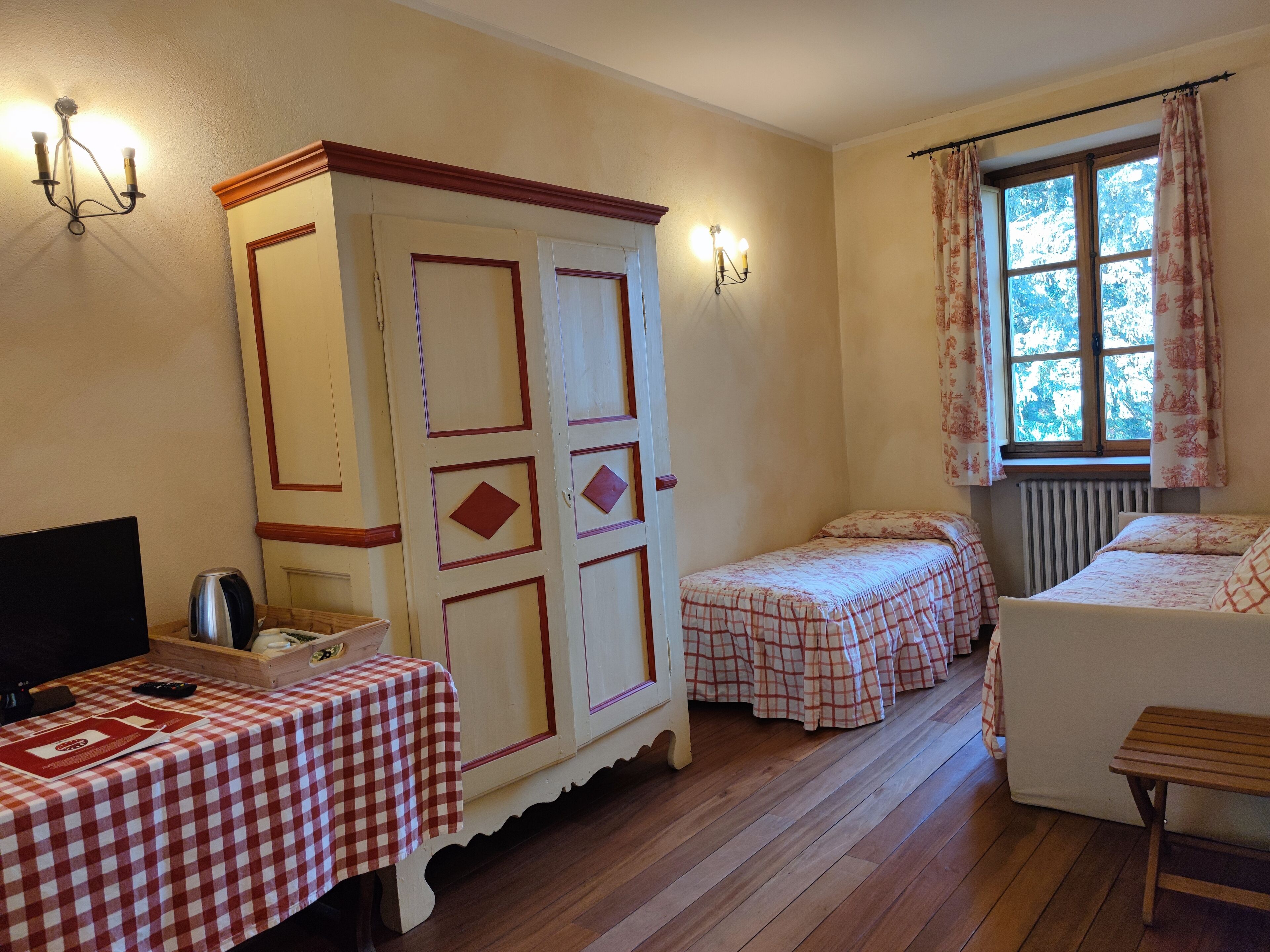 Una habitación con dos camas, una mesa de madera con mantel a cuadros, un armario y una ventana con cortinas.