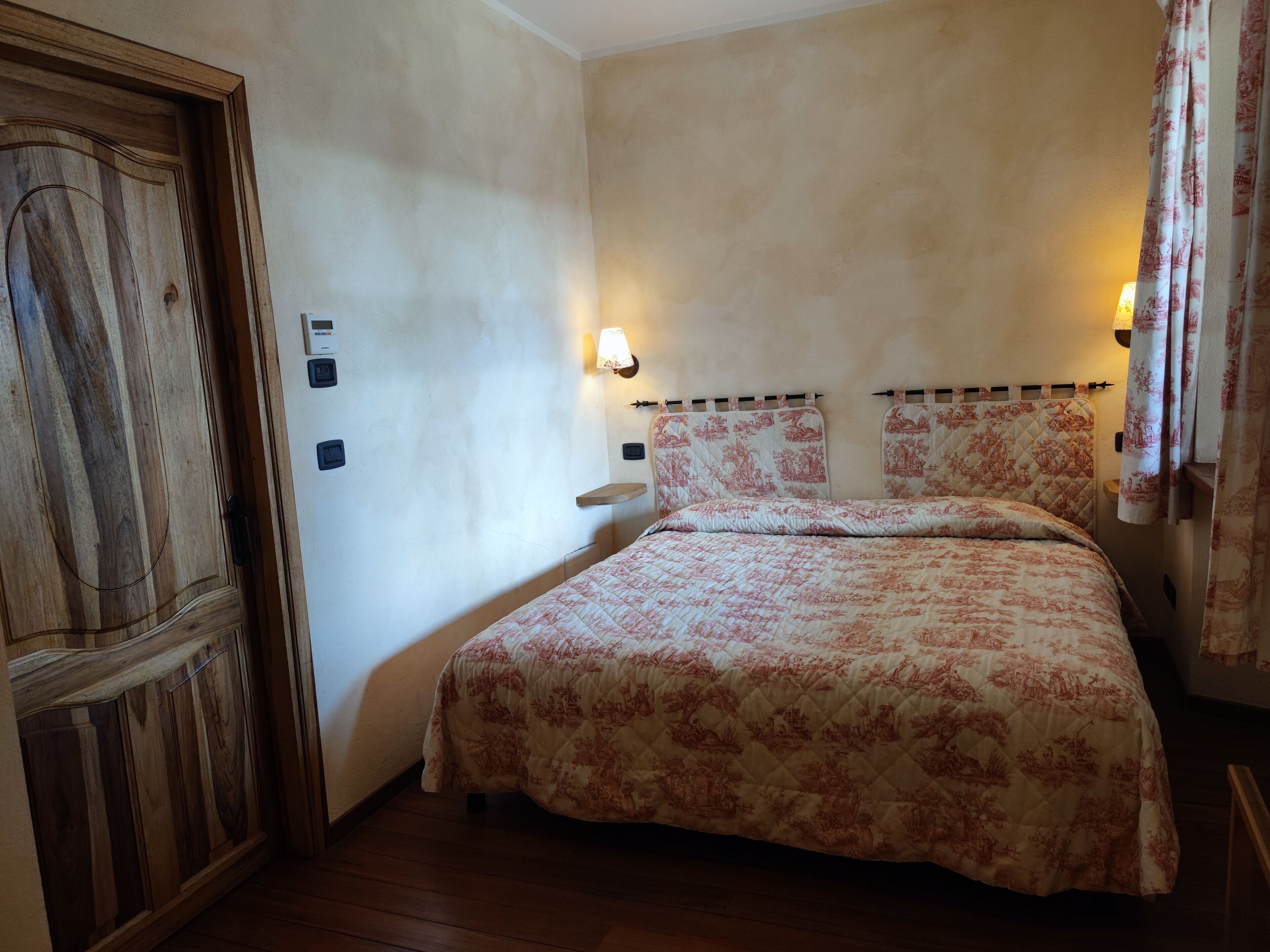 Une chambre avec une porte en bois, un lit recouvert d’une literie à motifs floraux, deux lampes fixées au mur et une petite étagère.
