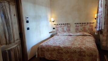 Une chambre avec une porte en bois, un lit recouvert d’une literie à motifs floraux, deux lampes fixées au mur et une petite étagère.